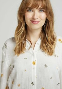 Chemise blanche à boutons en tissu texturé, ornée de broderies florales colorées en jaune, vert et blanc sur toute sa surface.