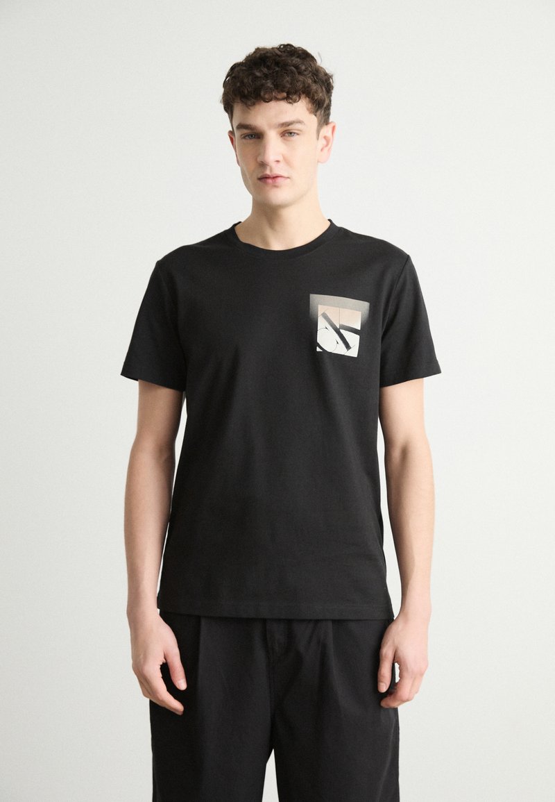 Camiseta negra de algodón con cuello redondo y mangas cortas. Presenta un bolsillo cuadrado con un gráfico geométrico en tonos neutros.