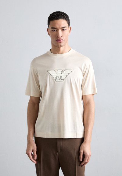 Crèmekleurig katoenen t-shirt met een groot geborduurd logo met vleugels in een gestructureerd ontwerp. Korte mouwen en een rechte snit.