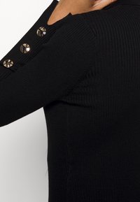 Černý žebrovaný top s dlouhým rukávem, kulatým výstřihem a třemi zlatými knoflíky na rameni, který se pyšní strukturovanou texturou a kontrastem.