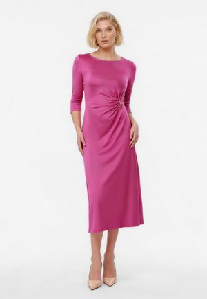 Mujer con cabello rubio corto, vistiendo un vestido magenta ajustado de manga larga y largo hasta la rodilla con fruncidos laterales y tacones puntiagudos beige.