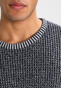 Dunkelblauer Strickpullover mit strukturiertem Finish, ausgestattet mit einem gestreiften Rippkragen. Eine Nahaufnahme hebt die Stoffdetails und das Muster hervor.