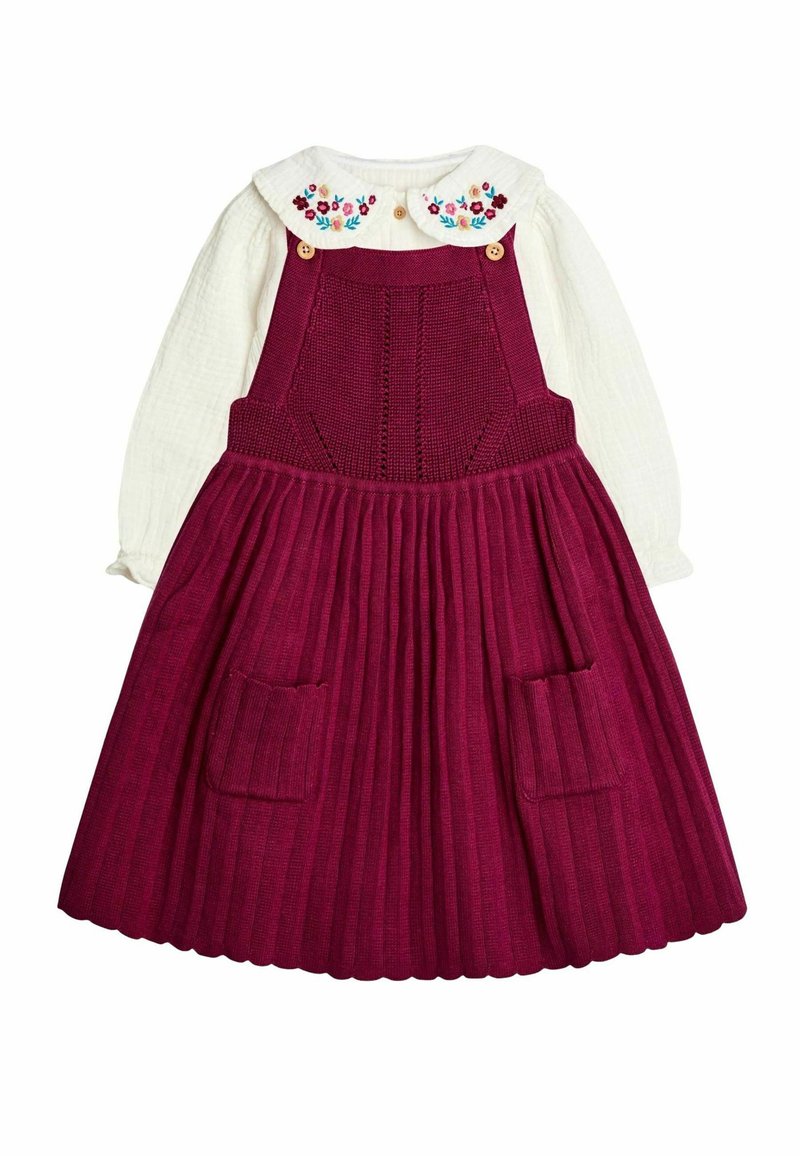 Weiße Bluse mit langen Ärmeln und besticktem Blumenkragen, kombiniert mit einem tiefroten Strickkleid mit Latz, das einen gefältelten Rock und praktische Fronttaschen aufweist.