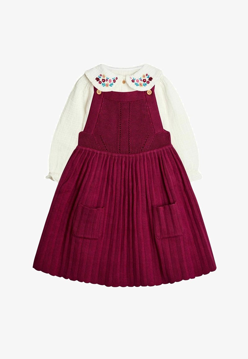 Weiße Bluse mit langen Ärmeln und besticktem Blumenkragen, kombiniert mit einem tiefroten Strickkleid mit Latz, das einen gefältelten Rock und praktische Fronttaschen aufweist.