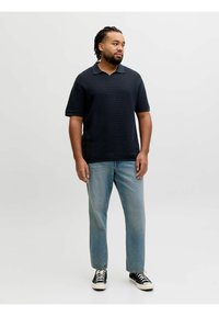 Homme aux cheveux tressés portant un polo noir texturé, un jean bleu clair et des baskets noires, debout devant un fond uni.