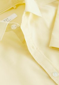 Camisa amarilla hecha de tejido texturizado con cuello puntiagudo, un botón y manga corta. La etiqueta dice "NEXT."