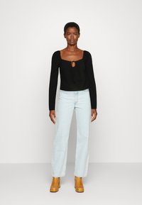 T-shirt preta de manga longa com decote quadrado e detalhe de laço à frente, combinada com jeans azul claro de perna reta e botas de salto castanhas.