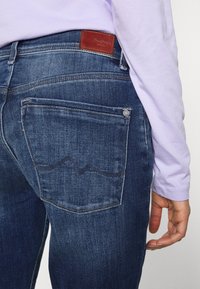 Jeans di denim blu scuro con un'etichetta in pelle sul retro, caratterizzati da cuciture in vista e un design con una sola tasca posteriore. Camicia a maniche lunghe viola morbida.