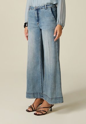 Kvinde iført lyseblå jeans med vide ben, lyseblå bluse med lange ærmer, sort armbånd og sorte strop-sandaler med hæl.