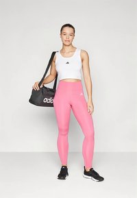 Top corto sin mangas blanco con logo negro, leggings rosas y zapatillas deportivas negras. Sosteniendo una bolsa de deporte negra con logo blanco.