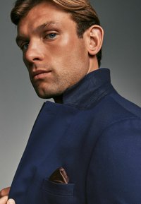 Marineblaue Jacke mit hohem Kragen, strukturiertem Stoff und einem braunen gemusterten Einstecktuch, das in die vordere Tasche gesteckt ist.