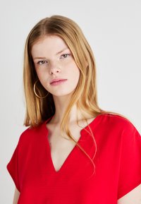 Maglietta rossa con scollatura a V, realizzata in tessuto testurizzato. Presenta maniche corte e una vestibilità comoda. Il modello indossa grandi orecchini a cerchio dorati.