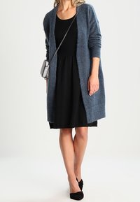 Donna che indossa un vestito nero e un cardigan lungo blu, porta una borsa a tracolla grigia e indossa tacchi neri, in piedi contro uno sfondo semplice.