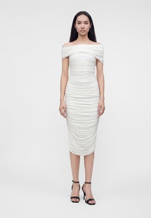 Vrouw met lang donker haar die een witte off-shoulder geplooide midijurk en zwarte hoge hakken sandalen draagt, staand tegen een effen achtergrond.
