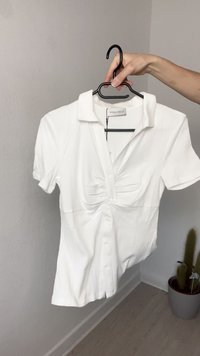 Camisa blanca de manga corta con cuello, que presenta plisados en el frente y cierre de botones. La tela parece acanalada y suave.