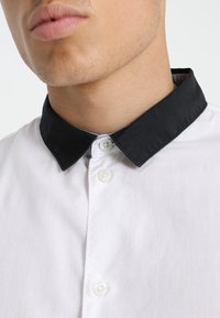 Camicia bianca con colletto nero, dotata di chiusure a bottone e una texture liscia. Focus sulla zona del collo e sul design del colletto.