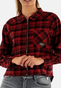 Donna che indossa una camicia a zip rossa e nera a scacchi con taschino sul petto e jeans blu sbiaditi, con le mani unite davanti.