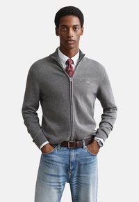 Muškarac u sivom džemperu na patentni zatvarač, bijeloj košulji, crvenoj kravati s uzorkom, plavim trapericama, smeđem remenu i ručnom satu s rukama u džepovima.