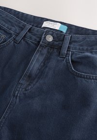 Tamsiai mėlynos spalvos džinsai su vidutinio aukščio liemeniu, tiesaus kirpimo, užsegami mygtukais, su priekinėmis kišenėmis. Etiketėje nurodyta „Tiesūs“ ir „Pagaminta Bangladeše“.