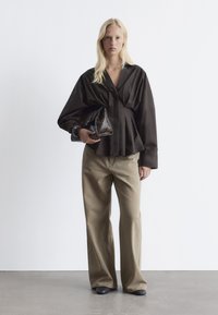 Dark brown, pleated blouse with a V-neck, wide sleeves; paired with light olive wide-leg pants and a textured clutch. Smooth fabric, casual style.