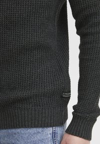 Ciemnoszary sweter dzianinowy z teksturowanym, waflowym wzorem oraz ściągaczami przy mankietach i dole. Mała metka marki na dolnym lewym boku.