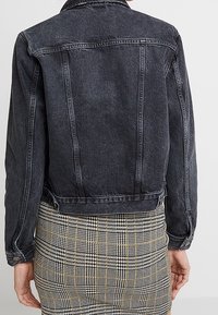 Zwarte spijkerjas met een aansluitend ontwerp, zichtbare naden en een iets verkorte lengte, gecombineerd met een geruite rok.