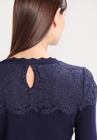 Mujer con cabello largo castaño que viste un top de punto azul marino con un yugo de encaje floral y una abertura en forma de lágrima en la parte posterior.