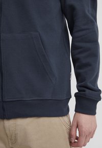 Tamno plava zip-up dukserica izrađena od mekog materijala, s prednjim džepom, rebrastim manžetama i malim logotipom sa strane.