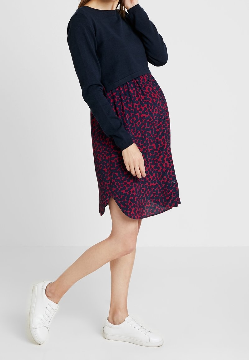 Femme portant un haut à manches longues bleu marine avec une jupe à pois rouges et bleus marins et des baskets blanches, debout avec une jambe légèrement levée.