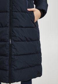 Main dans la poche avant d'une veste matelassée bleu navy avec une fermeture éclair visible et une patte de logo noire près de l'ourlet.