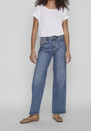 Kvinde iført højtaljede, vidde blå jeans, hvid t-shirt med op-rullede ærmer og beige balletsko, stående mod en grå baggrund.