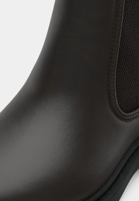 Hladká tmavě hnědá kožená Chelsea bota s elastickými bočními panely a černou podrážkou, zobrazená v detailním záběru.