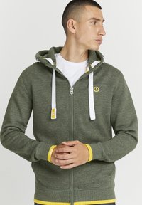 Green zip-up hoodie made from soft fabric, featuring white drawstrings, yellow accents, and a logo on the chest.