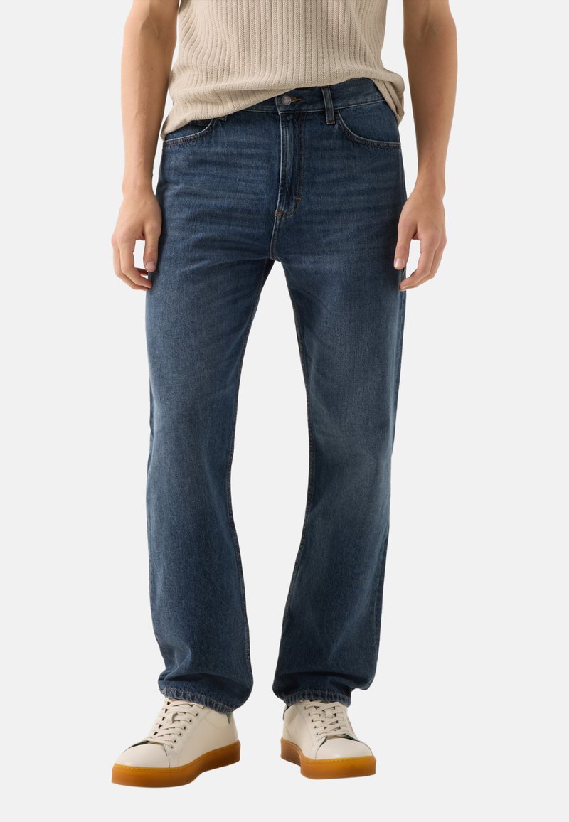 Person wearing relaxed-fit dark blue jeans, a beige ribbed sleeveless top, and white sneakers with gum soles.