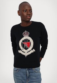 Zwarte gebreide trui met een groot schildlogo met een kroon, rode en witte accenten, en geribbelde mouwen en zoom. Ggedragen met denim jeans.