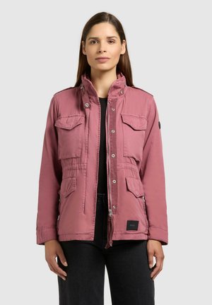 Femme portant une veste utilitaire rose poudré avec quatre poches à rabat à l'avant, sur un haut noir, debout devant un fond gris clair uni.