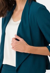 Morska marynarka z drapowanym przodem i rękawami o długości do łokcia, noszona na białej bluzce z srebrnym zdobieniem wzdłuż linii szyi.