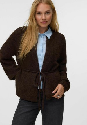 Femme blonde portant un cardigan marron foncé noué à la taille, par-dessus une chemise bleu clair boutonnée et un pantalon foncé, debout devant un fond uni.