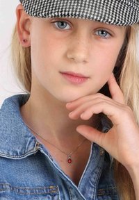 Fille portant une veste en denim avec un collier en chaîne argentée ornée d'un pendentif rouge et de petites boucles d'oreilles, sur un fond neutre.