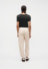 Άτομο με πλεγμένα μαλλιά στέκεται με την πλάτη γυρισμένη, φορώντας μαύρο μπλουζάκι, μπεζ παντελόνι τζόγκερ και ασπρόμαυρα αθλητικά σε λευκό φόντο.