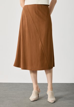 Brown A-line skirt made from soft fabric, featuring diagonal seam detailing. Paired with woven beige heeled mules on a gray surface.