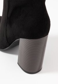 Stivaletto in suede nero con tacco grosso a strisce marroni. Lo stivaletto presenta una texture liscia e un design con la punta arrotondata.