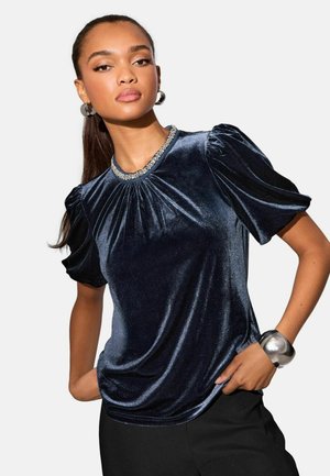 Marineblå velour-top med korte pufærmer, samlet halsudskæring og en perlekant. Glat tekstur, figursyet design, kombineret med et sølvarmbånd.