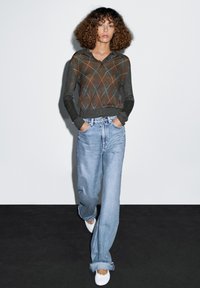 Donkergroene trui met een bruine ruitpatroon; losse, hoog gewaardeerde lichtblauwe jeans; witte schoenen; model dat tegen een effen achtergrond loopt.