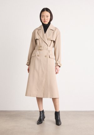 Cappotto beige con design doppiopetto, tasche anteriori, vita con cintura e ampi rever. Abbinato a stivaletti neri alla caviglia.