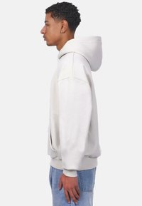 Hellgrauer Hoodie mit großer Kapuze, Kängurutasche und gerippten Bündchen. Hergestellt aus weichem Stoff mit lockerer Passform, getragen zu blauen Jeans.