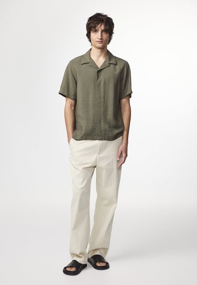 Camicetta verde oliva a maniche corte, pantaloni larghi a righe crema e sandali neri a infradito. Tessuto morbido con vestibilità rilassata e semplice design del colletto.