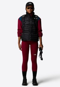 Black puffer vest, red fleece top, and burgundy leggings. Black cap, sunglasses, and durable shoes. Carrying trekking poles.