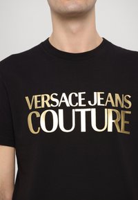 Juodas medvilnės marškinėliai su ryškiai auksine užrašų „VERSACE JEANS COUTURE“. Sklandus paviršius su klasikinio apvalaus kaklo dizainu.