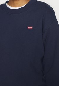 Tmavě modrá mikina z bavlněné směsi, s kulatým výstřihem a malým červeným logem Levi's na hrudi. Hladká textura.